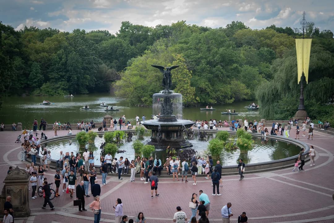 A large, ornate fountain anchors a busy park plaza, set against a backdrop of a pond and dense green forest.