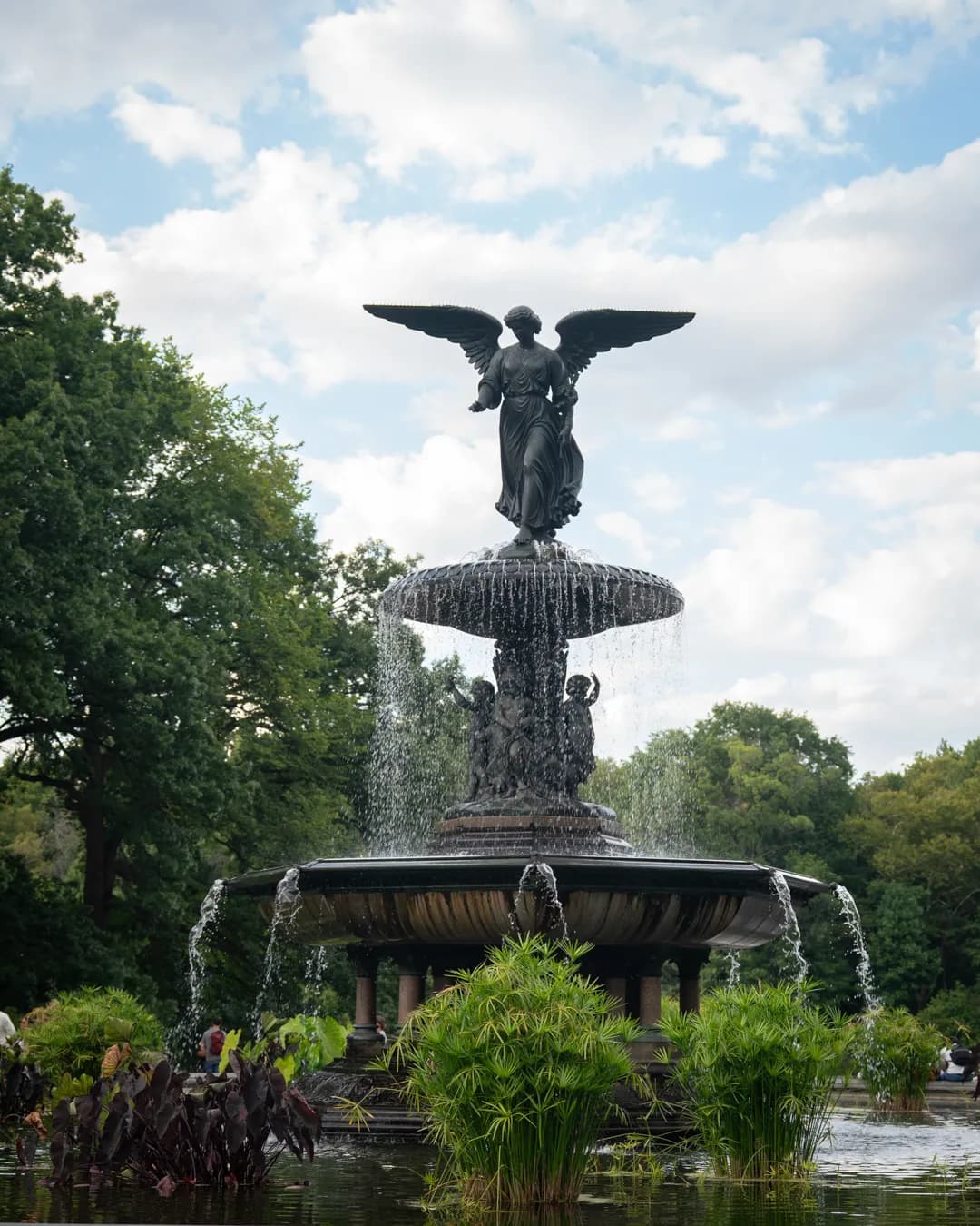 A grand, multi-tiered fountain featuring a winged statue sprays water in a lush park setting under a cloudy sky.