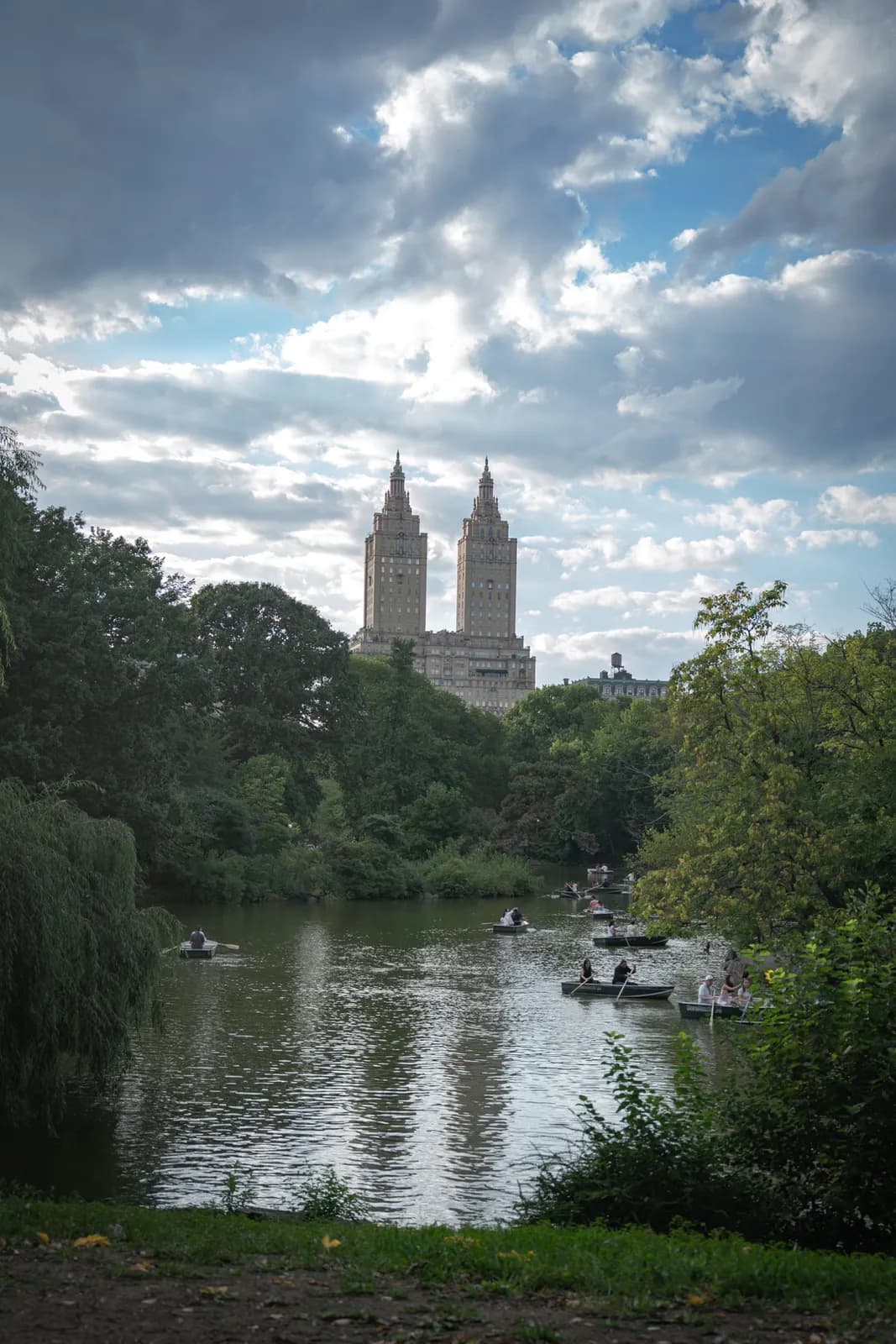 A scenic view of a river surrounded by dense foliage under a dramatic, cloudy sky, with prominent twin towers visible in the distance.