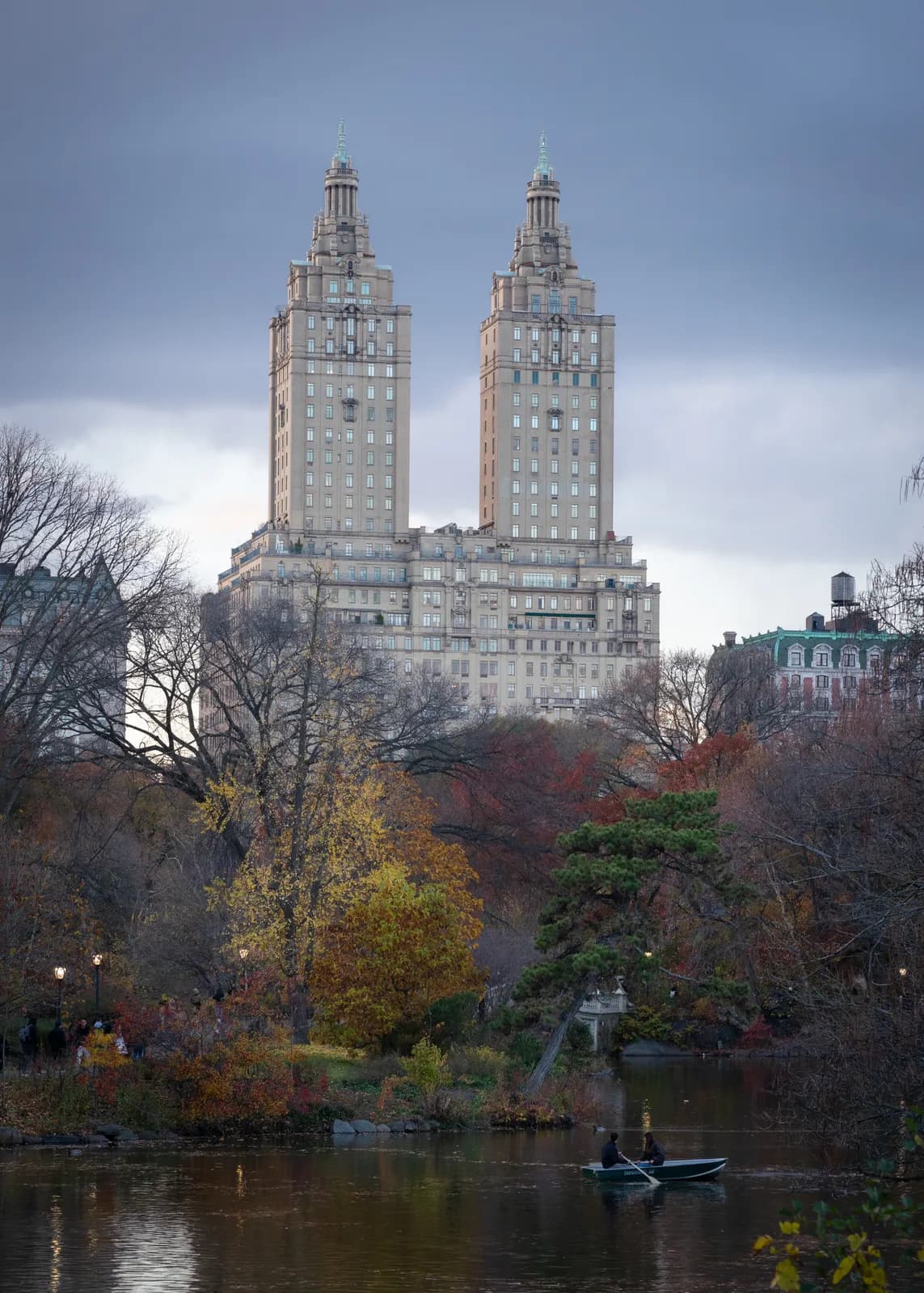 Two prominent skyscrapers dominate the skyline above a tranquil river scene framed by colorful autumn foliage under an overcast sky.